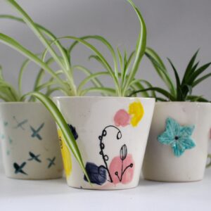 Trois petits cache-pots en céramique peints à la main, chacun avec des motifs uniques. Les pots contiennent des plantes vertes : l'un présente des dessins de petits oiseaux, le deuxième a des motifs colorés, et le troisième est décoré d'une fleur en relief. L'ensemble est placé sur un fond gris neutre.