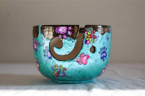 Un bol en céramique peint à la main, décoré de motifs colorés de pattes et de fleurs sur un fond turquoise éclatant. Le design unique présente une finition brillante et une découpe stylisée sur le bord. Cette pièce artisanale est posée sur un fond neutre, permettant de mettre en valeur ses couleurs vives et son charme ludique.